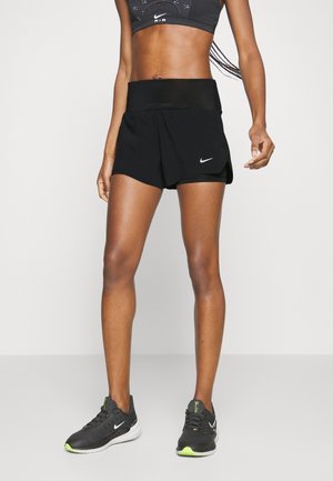Kobieta ubrana w czarny sportowy biustonosz Nike, czarne spodenki do biegania Nike oraz czarne buty do biegania Nike z neonowo zielonymi akcentami, stojąca na szarej podłodze.