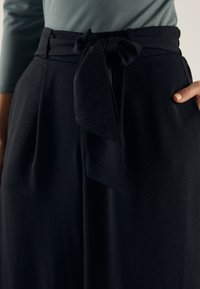 Dark navy pleated trousers with a self-tie belt at the waist, featuring soft, textured fabric and side pockets.