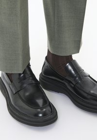 Mocasines de cuero negro con un acabado brillante, una punta ligeramente afilada y una suela de goma gruesa. Combinados con pantalones verdes a medida y calcetines marrones.