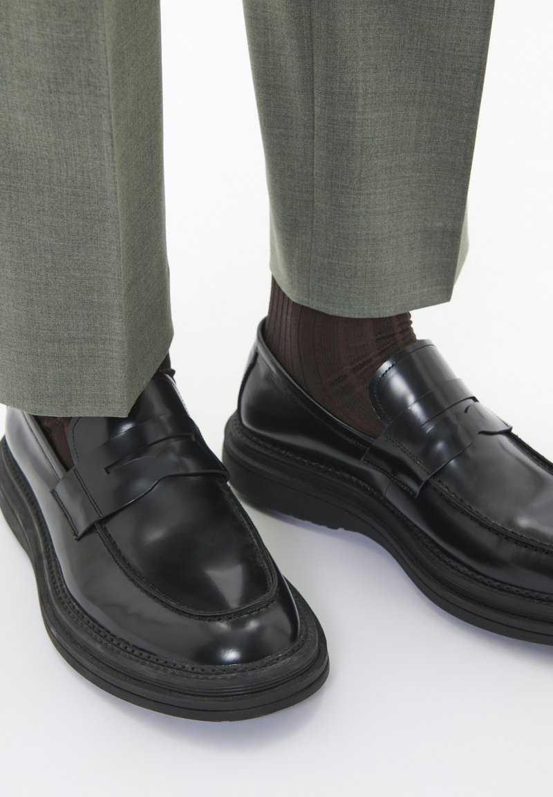 Mocasines de cuero negro con un acabado brillante, una punta ligeramente afilada y una suela de goma gruesa. Combinados con pantalones verdes a medida y calcetines marrones.