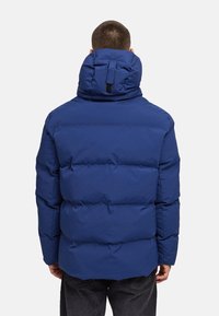 Modrá prešívaná bunda s vysokým golierom a nastaviteľnou kapucňou, s výrazným prešívaním a zaujímavými detailmi.