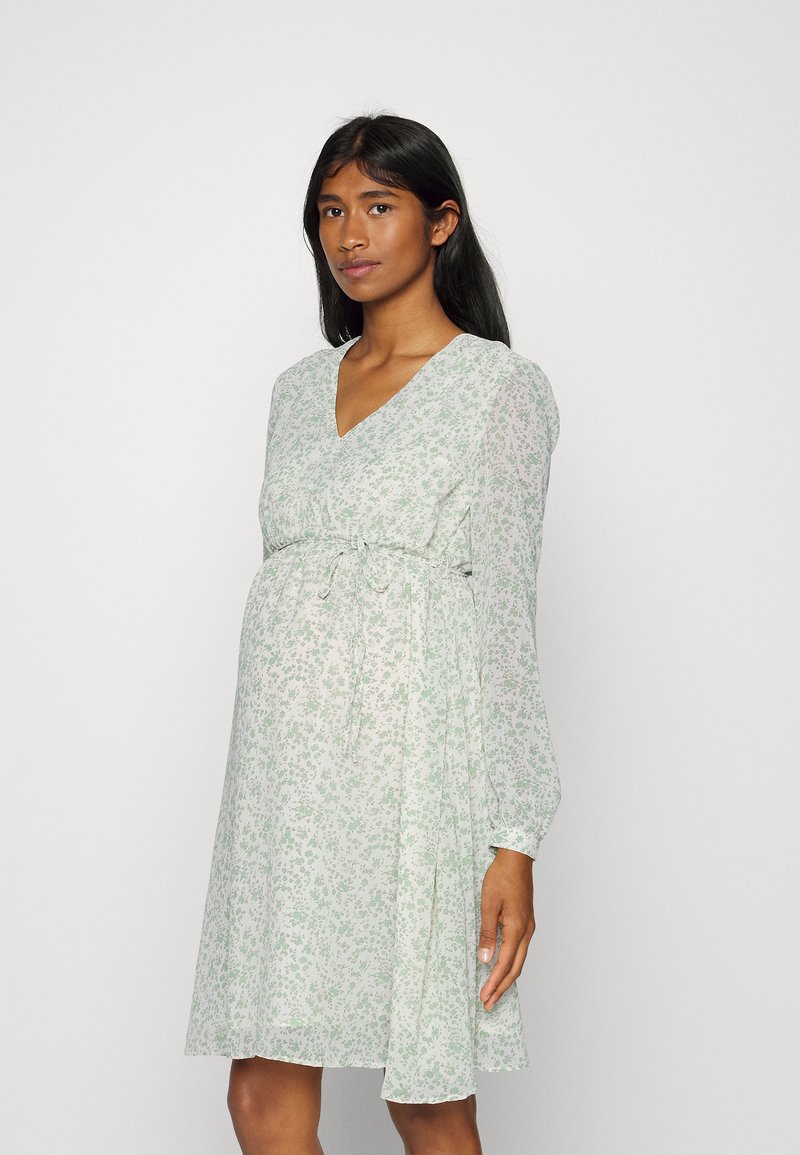 Vestido de maternidade floral verde claro com decote em V, mangas longas, cintura ajustável e saia fluida, apresentando uma textura de tecido suave e transparente.