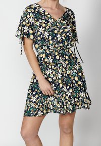 Žena v krátkých námořnických modrých šatech s květinovým vzorem a volánkovým lemem, které mají krátké rukávy vázané mašličkami, na jednobarevném světlém pozadí.