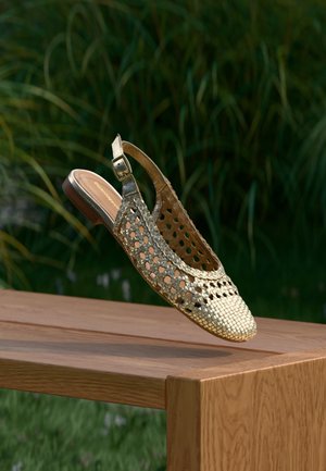 Scarpa slingback in pelle intrecciata oro metallico con cinturino e fibbia, sospesa sopra una panchina di legno all'aperto con vegetazione sullo sfondo.