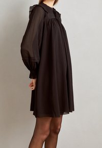 Robe en chiffon noir avec des manches longues, un col froncé et des accents en volants. Silhouette fluide avec des poignets élastiques et une texture transparente.