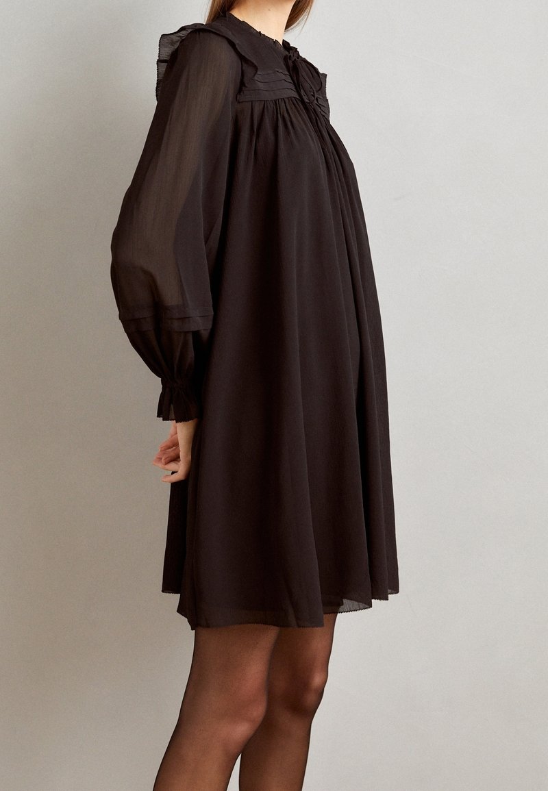 Robe en chiffon noir avec des manches longues, un col froncé et des accents en volants. Silhouette fluide avec des poignets élastiques et une texture transparente.