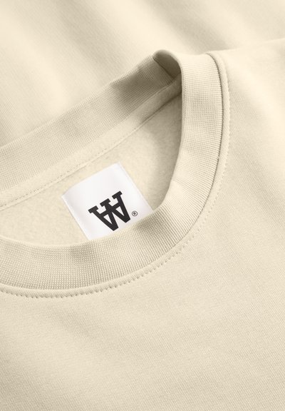 Sweatshirt beige avec un col rond côtelé et une étiquette en tissu avec un logo stylisé. Texture lisse, design minimaliste.