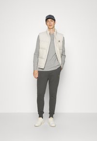 Lichtbeige gewatteerde vest over een grijs langemouwen shirt, gecombineerd met donkergrijze joggingbroeken met een geel logo, en witte sneakers.