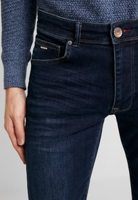 Jeans de mezclilla azul oscuro, con un diseño clásico de cinco bolsillos, costuras visibles y un botón de marca cerca de la cintura.