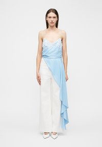 Νέα γυναίκα με ίσια καστανά μαλλιά, φοράει ανοιχτόγαλανη καμιζόλα με δαντέλα και ριχτό ύφασμα, συνδυασμένη με λευκό παντελόνι φαρδιάς γραμμής, στέκεται ακίνητη.