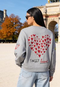 Femme portant un sweat gris avec des cœurs rouges formant un grand cœur et le texte "SWEET heart", debout à l’extérieur près d’un monument en forme d’arche.