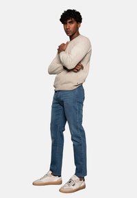 Suéter de punto beige, jeans azules y zapatillas ligeras con una lengüeta oscura. La persona tiene los brazos cruzados, con un fondo neutro.