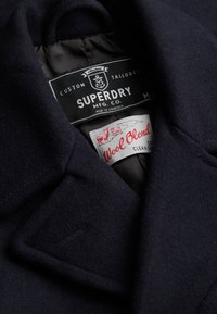 Marineblauwe jas van wolmix met Superdry-merklabel en onderhoudsetiket van wolmix aan de binnenkant van de kraag.