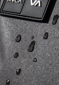 Tejido gris con una superficie texturizada que muestra múltiples gotas de agua. Presenta la marca RVCA en material negro gomoso.