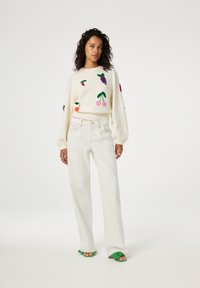 Witte trui met kleurrijke fruitborduursels; oversized pasvorm en geribbelde manchetten. Gecombineerd met witte jeans met wijde pijpen en groene slippers.