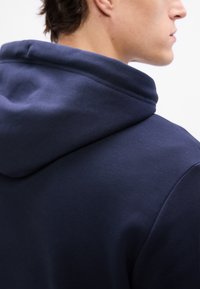 Námornícky modrá mikina s kapucňou s rebrovanou textúrou. Kapucňa je mierne prehnaná a disponuje šitým švom na zadnej strane.