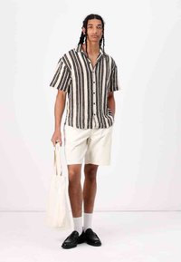 Camisa de botones de rayas blancas y negras de manga corta combinada con pantalones cortos claros, calcetines blancos, mocasines negros y un bolso de mano de color crema.