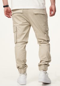 Beige Cargo-Hosen mit elastischen Bündchen und mehreren Seitentaschen, kombiniert mit weißen Sneakers. Hergestellt aus einem glatten, leichten Material.