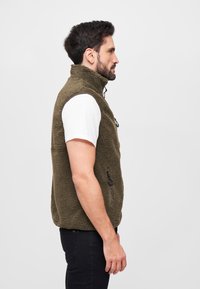Fleece-Weste in Olivgrün mit hohem Kragen, seitlicher Reißverschlusstasche und strukturierter Oberfläche. Über einem weißen T-Shirt getragen.