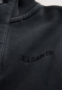 Schwarzer Strickstoff mit geripptem Kragen, der schwarzen bestickten ALLSAINTS Logo-Schriftzug auf dem Brustbereich zeigt.