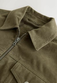 Alyvuogių žalios spalvos zomšinė striukė su sulankstytu apykakliu, matoma sidabro spalvos užtrauktukas iš dalies užsegta ir siūta kišenės sklendė.