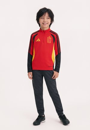 Chlapec stojaci v červeno-žlto-čiernej športovej bunde s erbom, čiernych nohaviciach a topánkach, usmievajúci sa a pozerajúci doprava.