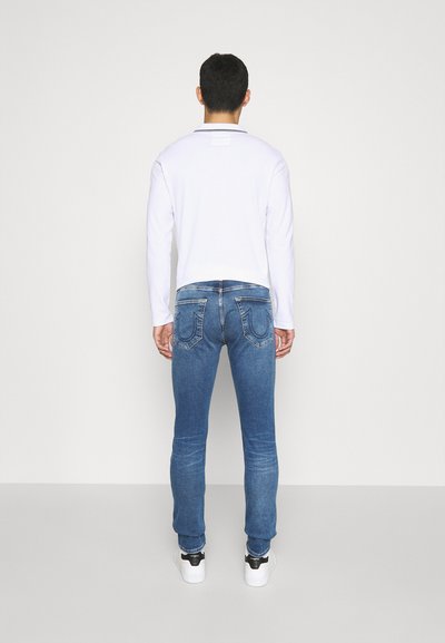 Blaue Jeans im Slim Fit, mit zwei hinteren Taschen und dekorativen Nähten. Kombiniert mit einem weißen Langarmhemd und Turnschuhen.