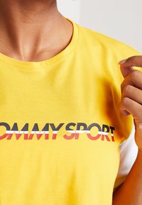 Žluté tričko s krátkým rukávem vyrobené z bavlny, s potiskem "TOMMY SPORT" v tučných černých, červených a bílých písmenech přes hruď.