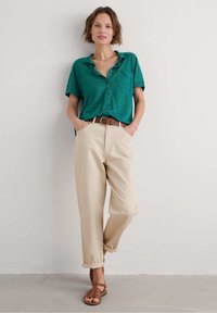 Green short-sleeve button-up shirt, beige wide-leg trousers with rolled cuffs, brown leather belt, and brown strappy sandals.