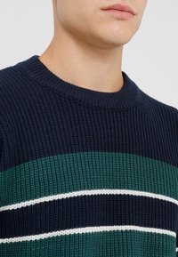 Pull en maille bleu marine avec des rayures horizontales vertes et blanches. Présente une texture côtelée et un col rond. Vue en gros plan.