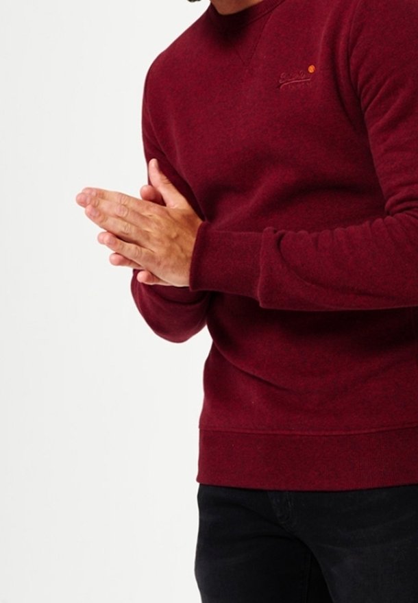 Homme portant un pull à manches longues bordeaux, mains croisées sur un fond blanc uni.