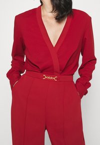 Mono rojo ajustado con un escote en V profundo, mangas largas y un cinturón a juego con detalles de hebilla dorada, diseñado con una tela suave.