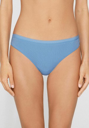 Frau trägt hellblaue Bikini-Unterwäsche, gezeigt vom mittleren Oberkörper bis zu den oberen Oberschenkeln vor einfarbigem Hintergrund.