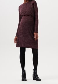 Långärmad, vinröd klänning i en mjuk trikå med draperad midja. Mjukt stickat material, kortare än låret, kombinerat med svarta ankelboots och strumpbyxor.