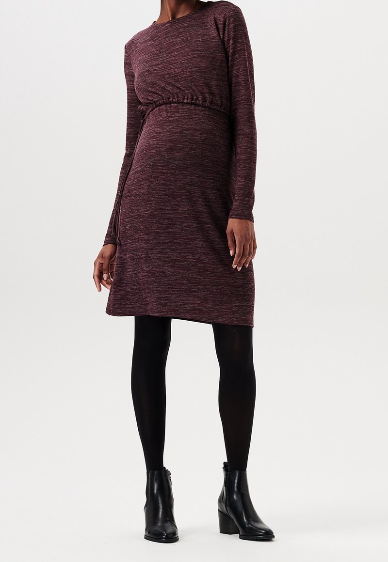 Långärmad, vinröd klänning i en mjuk trikå med draperad midja. Mjukt stickat material, kortare än låret, kombinerat med svarta ankelboots och strumpbyxor.