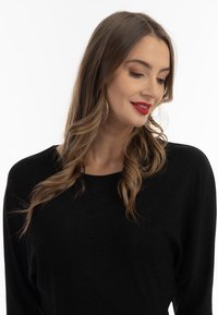 Czarny longsleeve z luźnym krojem i okrągłym dekoltem. Gładka, miękka faktura. Modelka nosi duże kolczyki i jasną czerwoną szminkę.