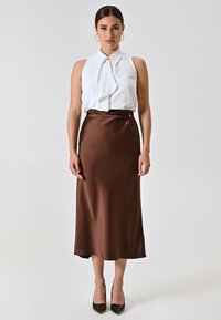 Blusa bianca di raso con una parte anteriore e colletto arricciati, abbinata a una gonna setosa marrone scuro al ginocchio e décolleté neri a punta.