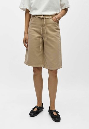 Person iført beige knælange shorts med bindebånd foran, hvid kortærmet top, sorte Mary Jane-sko og et sølvarmbånd på venstre håndled.