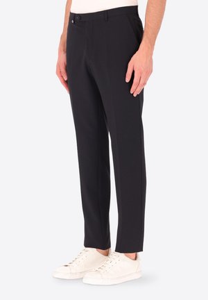 Uomo che indossa pantaloni neri slim fit e sneakers bianche, in piedi con le mani rilassate lungo i fianchi su uno sfondo bianco semplice.