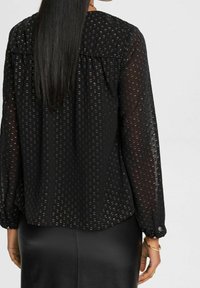 Femme aux longs cheveux noirs raides portant un chemisier noir transparent à petits points en relief et une jupe en cuir noire, vue de dos.