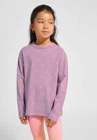 Junges Mädchen mit langen dunklen Haaren, das ein lila Langarmshirt mit gelben Mustern und hellrosa Leggings trägt und zur Seite schaut.