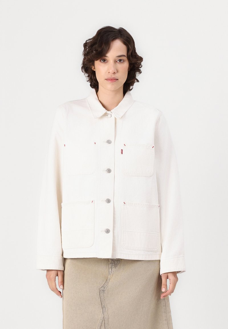 Veste en denim blanche avec une fermeture à boutons, coupe oversize et deux poches avant ; présente des boutons argentés et des accents rouges.