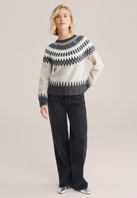 Sweter robiony na drutach w kolorze kremowym i ciemnoszarym z geometrycznym wzorem. Posiada okrągły dekolt oraz długie rękawy; zestawiony z czarnymi spodniami o szerokich nogawkach.