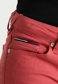 Jeans de mezclilla rojos con cierre de cremallera y botón, que presentan un pequeño bolsillo con un patrón tricolor en el interior y acentos metálicos.