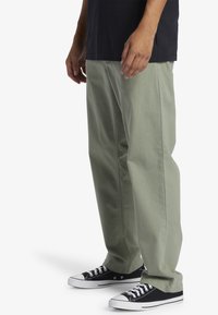 Pantaloni verzi cu picioare largi, din bumbac, cu o textură solidă, buzunare laterale și asortați cu adidași negri cu tălpi albe.