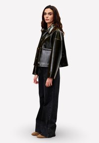 Vrouw met golvend bruin haar draagt een glanzende zwarte oversized jas, een brede zwarte jeans en bruine puntige schoenen, en staat zijwaarts op een witte achtergrond.