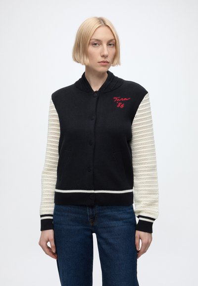 Femme aux cheveux blonds courts portant un pull noir boutonné avec des manches en crochet blanc et un jean bleu, faisant face.