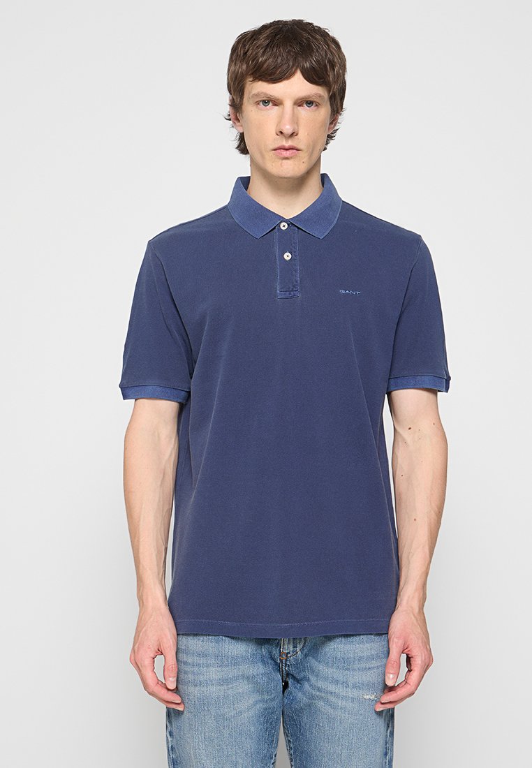 Mężczyzna w gładkiej granatowej koszulce polo i jasnoniebieskich dżinsach, stojący na jasnoszarym tle.