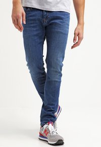 Blå denimjeans med slim fit, som visar en mjuk textur och minimal sömnad, tillsammans med grå och röda sportskor.