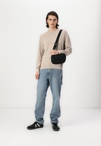 Borsa a tracolla nera con dettagli in zip indossata da una persona con un maglione beige e jeans azzurri, in piedi su uno sfondo bianco.